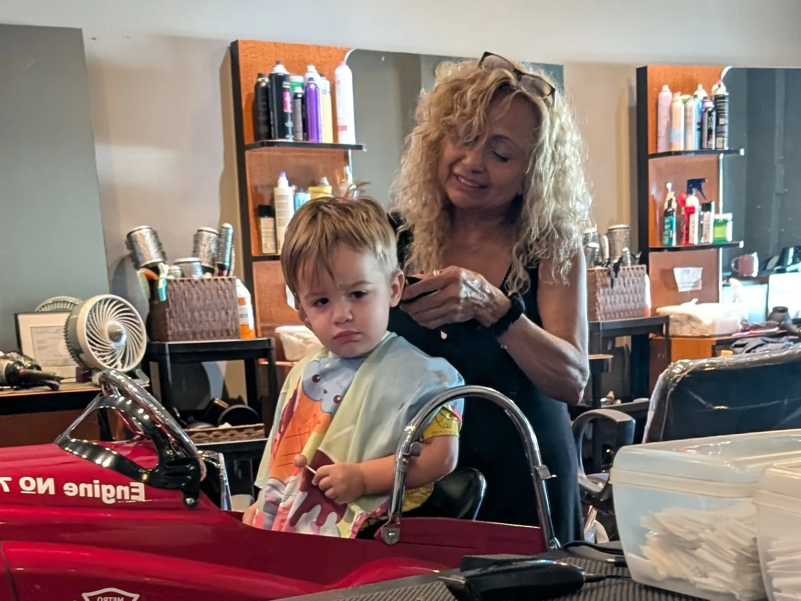 A stylist trims a toddler’s hair in the fire truck chair, in front of wooden shelves of hair products
