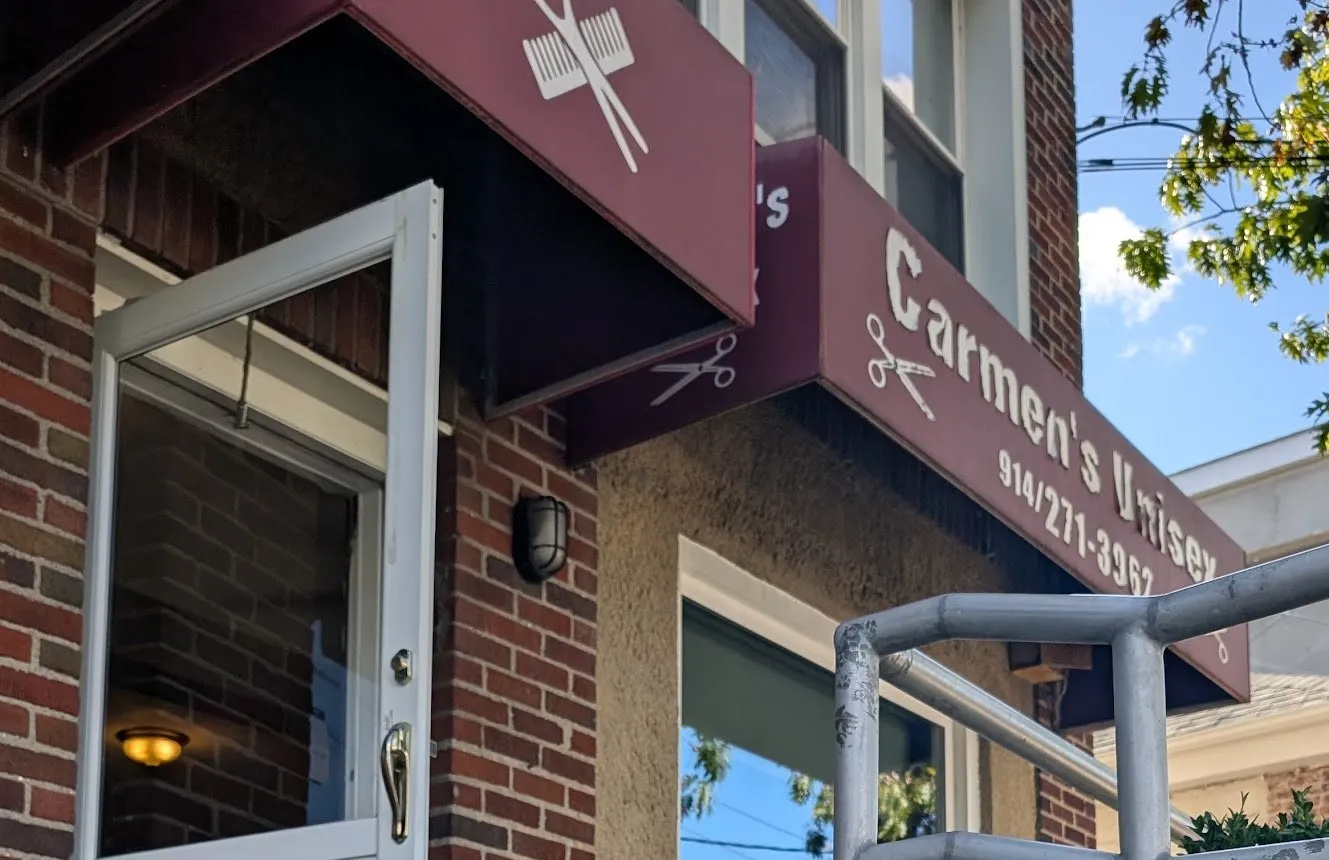 The burgundy awning of Carmen’s Unisex, with white scissors and the phone number, over a red-brick storefront and a white door