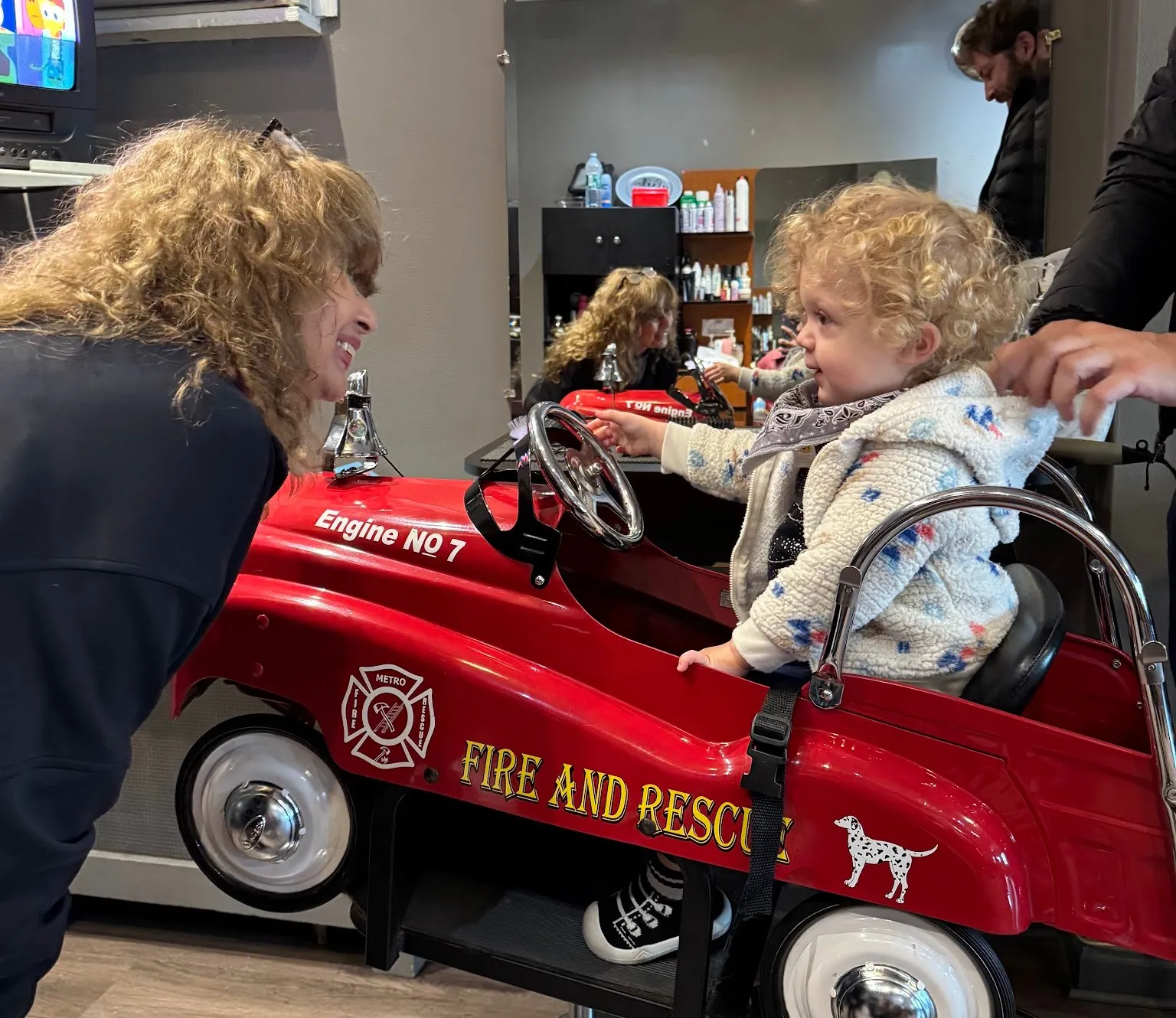 A stylist smiles at a curly-haired toddler sitting in the red fire truck chair at Carmen’s Unisex