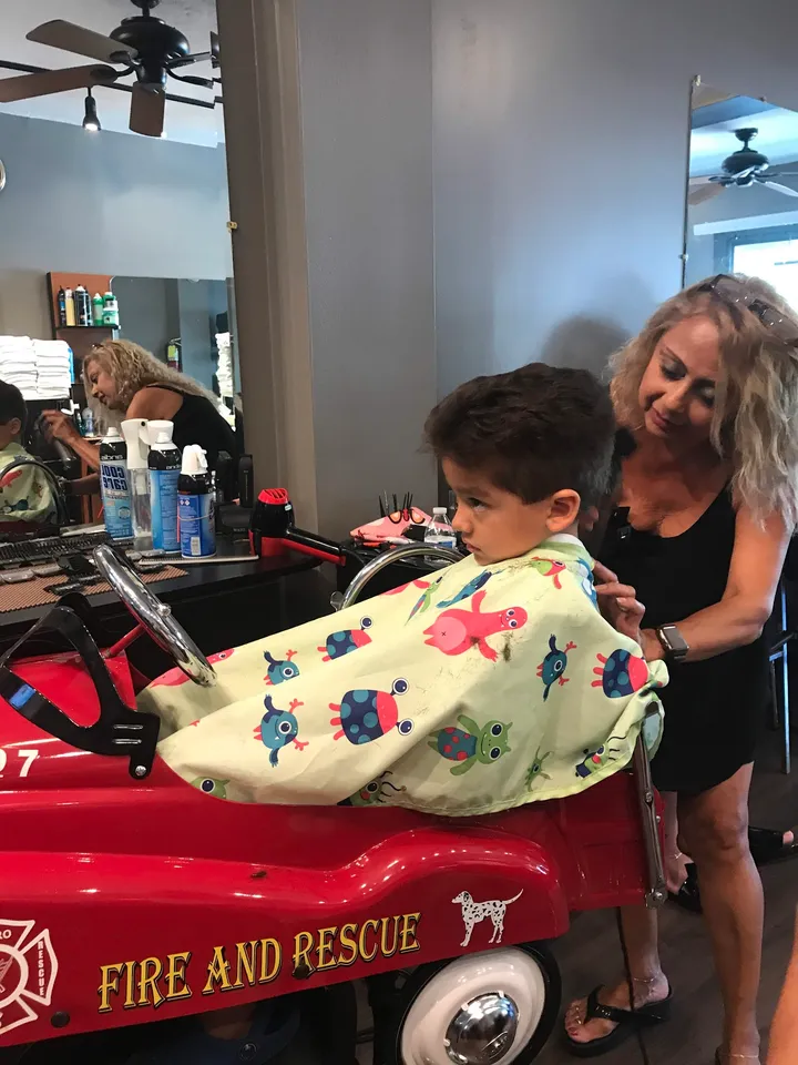 A stylist trims a boy’s short haircut while he sits in the fire truck chair in a printed cape