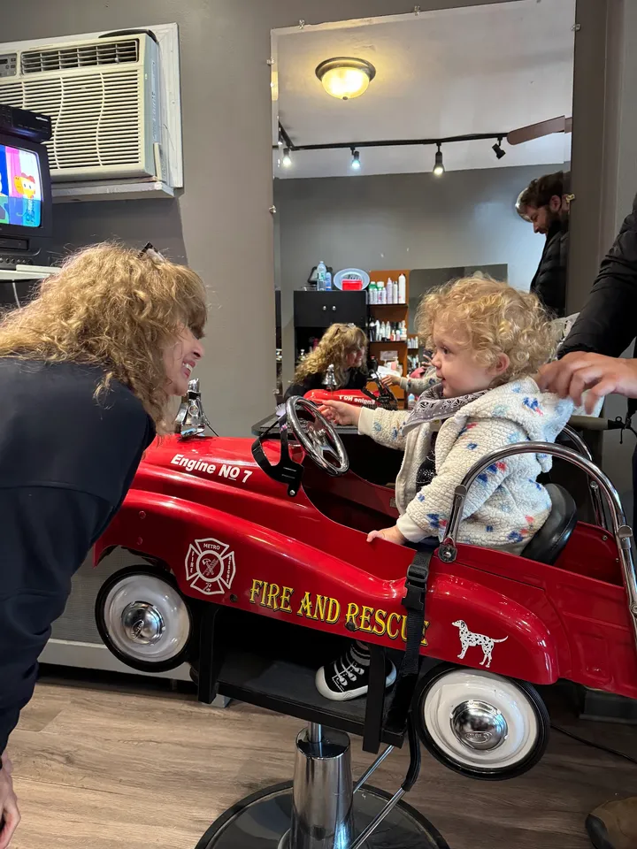 A curly-haired toddler at the wheel of the fire truck chair, smiling back at the stylist