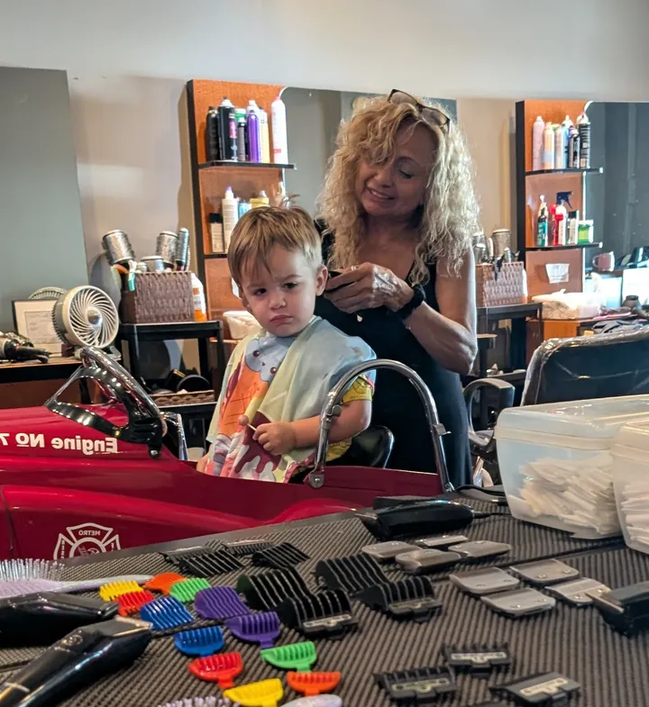 A stylist trims a toddler’s hair in the fire truck chair, with clipper guards in every color on the station