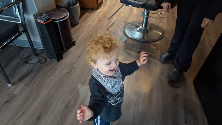 A curly-haired toddler walks across the wood floor after a first haircut