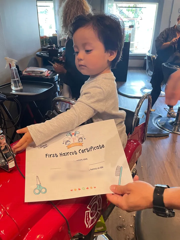 A toddler with a fresh haircut and a First Haircut Certificate, sitting in the fire truck chair