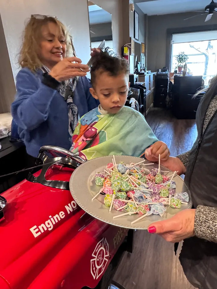 A child in the fire truck chair picks a lollipop from a full tray after a haircut